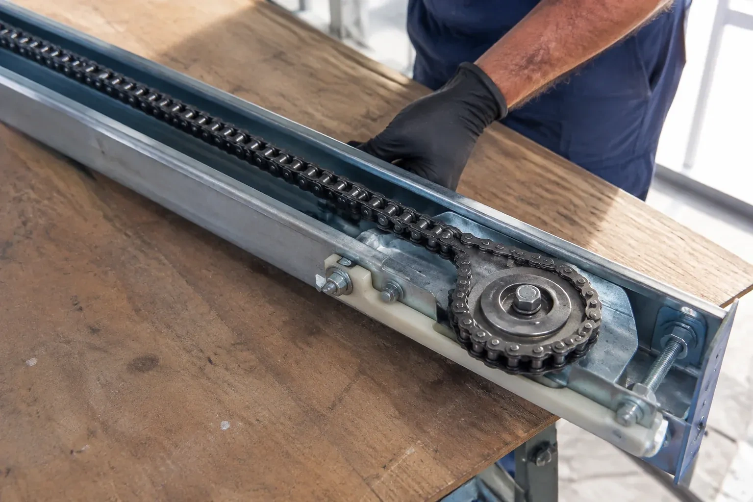 Gloved hand resting on a steel opener rail laid on a wooden bench, showing a drive chain looped around a sprocket wheel. — Chain-Drive Opener Repair, London