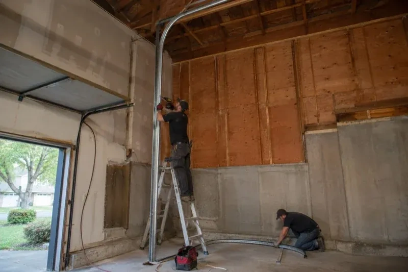 One person on a stepladder working beside a tall curved garage-door track while another kneels beside loose track sections in an unfinished garage. — High-Lift Garage Door Conversion, London